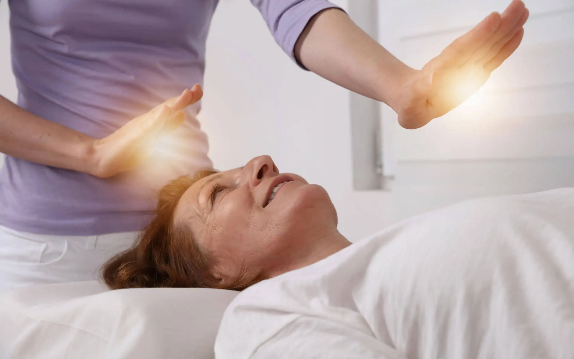 A smiling woman lying on a massage table receiving healing energy from another woman's hands